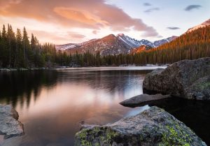 Photo of Bear Lake in Rocky Mountain National Park, Colorado at sunrise. Pink sky with the sheer flanks of Hallett Peak in the distance framed by evergreens, mirrored in the glass-like lake.